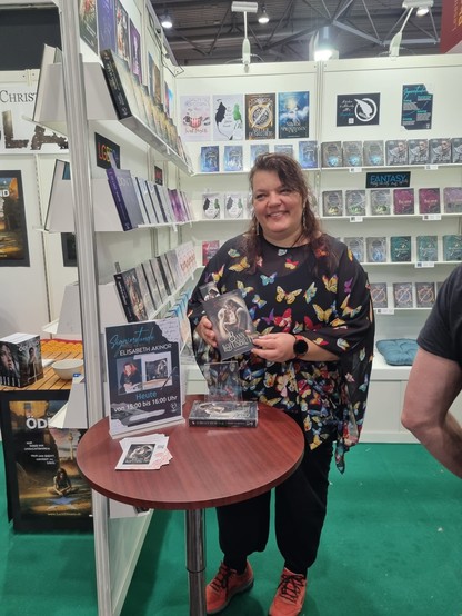 Eine Frau steht an einem Stand auf der Leipziger Buchmesse. Sie hat lange braune Haare und trägt eine schwarze Tunika mit Schmetterlingen bedruckt. In der Hand hält sie ihren Debutroman 