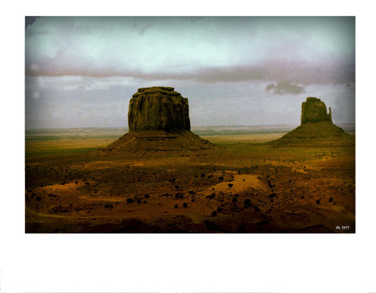Old color photo (1977) from Monument Valley, Arizona, showing two of the stone monuments rising from the desert. The landscape towards the horizon is greener. Moody grey sky.
