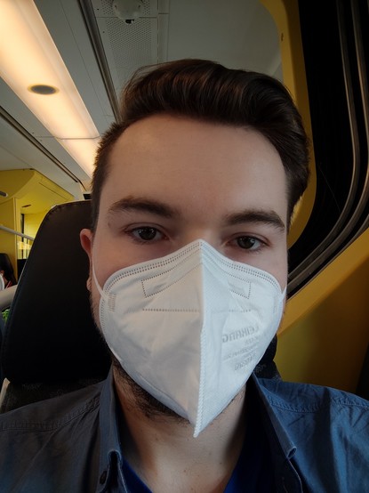 27 years old white cis man with short brown hair sitting on the train to Karlsruhe, wearing an FFP2 mask and a blue shirt.