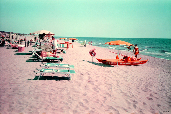 Farbfoto, das einen Ausschnitt des Strands in Ostia am Mittelmeer zeigt. Im Vordergrund rosa Sand. Links leere und benutzte Liegen, rechts ein oranges Rettungsboot (Katamaran) mit Sonnenschirm. Rechts das türkise Meer. Hell türkiser Himmel. Verfremdete Farben wegen des verwendeten Purple-Films.