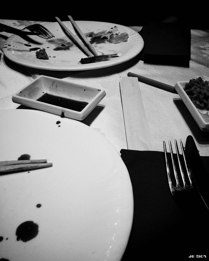 Schwarzweißfoto, das die Überreste einer Sushi-Mahlzeit auf einem Tisch zeigt. Teller mit Eßstäbchen und Tropfen von Sojasoße unten links, in Bildmitte eine Schale mit übriger Sojasoße, oben ein weiterer Teller mit Speiseresten, Eßstäbchen und Gabel. Rechts unten eine dunkle Serviette mit Gabel darauf, darüber eine Schale mit Speiseresten.