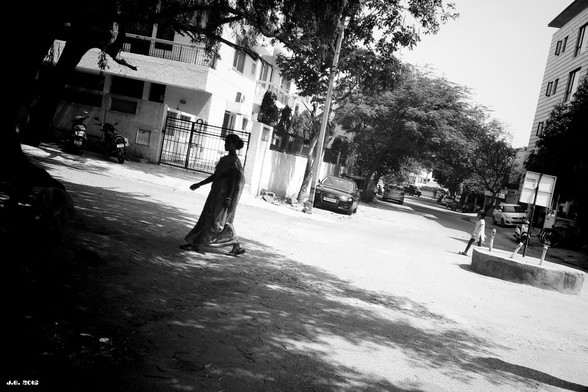 Schwarzweißfoto einer Straßenszene in New Delhi. Erfundene Bildbeschreibung: Mrs. Chatterjee auf dem Nachhauseweg nach dem Kauf dreier Äpfel. Ihr gerade in Ungnade gefallener Sohn Saurab folgt rechts in sicherem Abstand.