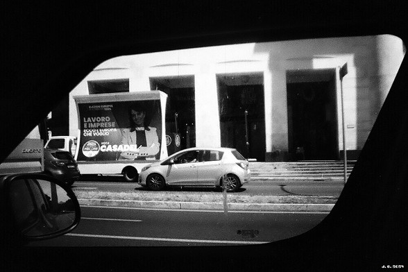 Schwarzweißfoto - aus dem stehenden Auto durchs Seitenfenster aufgenommen. Blick auf eine Straße mit einem Kleinwagen, einem Lkw mit Werbewand. Dahinter ein Geschäftsgebäude, zu dessen Eingang eine flache Treppe hochführt.