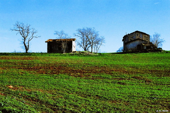 Farbfoto - ländliche Gegend in Lazio außerhalb von Rom. Im Vordergrund ein ansteigendes Feld mit spärlichem Grün auf brauner Erde. Am Horizont ein verfallener Bauernhof, der aus einem Haupt- und einem Nebengebäude besteht, und etliche dürre Laubbäume unter blauem Himmel.