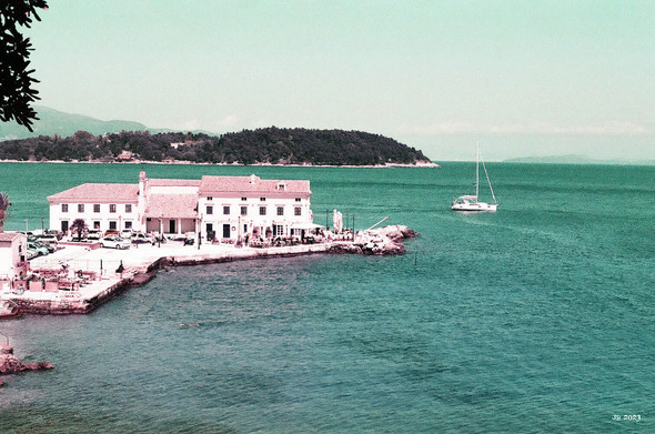 Farbfoto - Blick auf die Adria in Richtung Albanien von Korfu aus. Im Vordergrund das türkise Meer, dann ein großes weißes Gebäude mit Landeplatz, ein vertäutes Segelboot, weiter entfernt Teile einer Insel mit grünem Bewuchs. Verfremdete Farben durch den verwendeten Farb-Infrarotfilm. Olympus XA auf Lomochrome Purple.