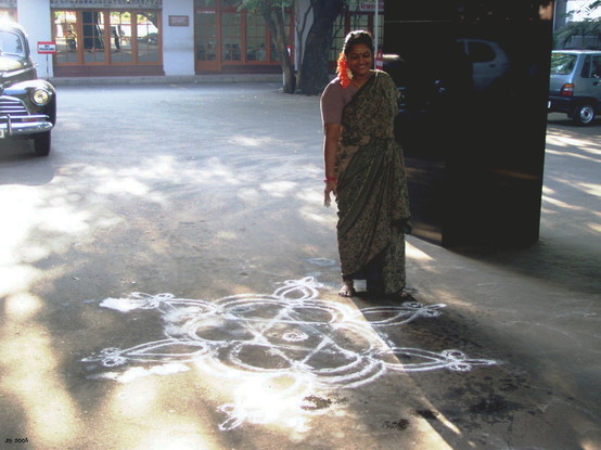 Farbfoto - Eingangsbereich eines Hotels in Chennai, Indien. Auf dem Betonboden ein Kolam - ein symmetrisches Muster aus Reismehl, das jeden Tag neu gemalt wird - und dahinter dessen Urheberin, eine junge Frau in traditionaller Kleidung. Ihre Fingerspitzen sind weiß von dem Mehl.