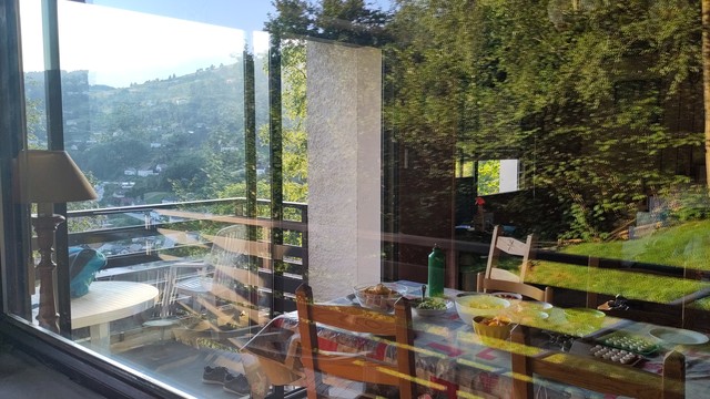 Doppelte Reflexion durch zwei Fensterfronten, auf dem Bild eine grüne Bergwiese und Büsche in der Abendsonne, ein innen gedeckter Tisch mit Tellern und vielen Schalen, und eine dunkle Bergsilhouette mit vielen Chalets als helle Punkte.