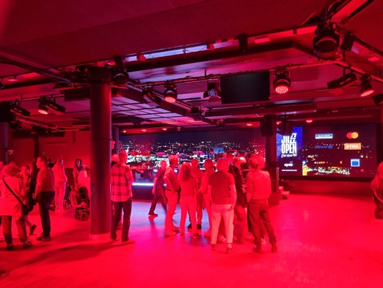 Menschen im roten Scheinwerferlicht stehend, dahinter eine Bühne und rechts daneben eine Leuchtreklame mit dem JazzOpen-Schriftzug und den Logos der Sponsoren.