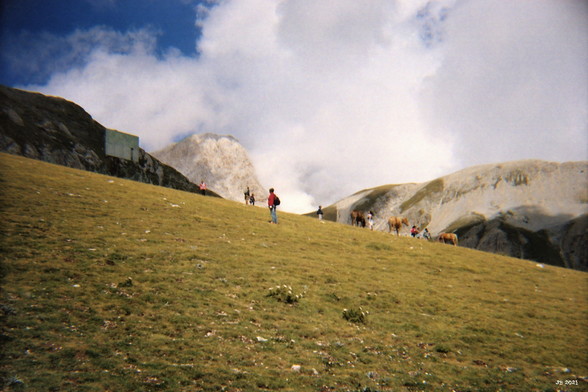 Farbfoto der Berglandschaft unterhalb des Gran Sasso in den Abruzzen in Italien. Im Vordergrund eine grüne Wiese mit vereinzelten Steinen, in Bildmitte etliche Wanderer und Pferde, darüber die hohen Berge, mit der Spitze des Gran Sasso in der Mitte. Am Horizont blauer Himmel mit großen weißen Wolken.