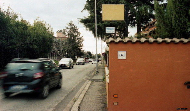 Farbfoto, das rechts eine braun-orange Mauer mit gewellter Ziegelabdeckung und der Zahl 261 auf einer weißen Kachel zeigt, darüber grüne Bäume, Schilder und ein Teil eines Hauses. Auf der linken Seite eine befahrene Durchgangsstraße mit etlichen Autos. Das schwarze Auto vorne links ist sichtbar in Bewegung. Ganz links grüne Bäume. Blassblauer Himmel über allem.