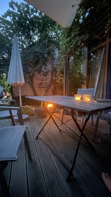 Ein hölzerner Terrassentisch mit Kerzen darauf im Hintergrund noch mal Kerzen, ein Feigenbaum und eine begrünte Hausfassade