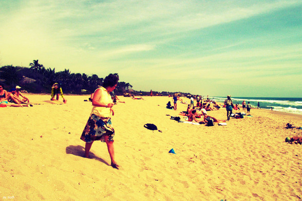 Farbfoto vom Strand von Auroville in Tamil Nadu, Südindien. In der unteren Bildhälfte viel gelber Sand. Im Vordergrund eine Frau unterwegs zum Wasser. Über den Strand verteilt Leute auf Handtüchern oder stehend. Am Horizont links grüner Wald, rechts das Meer mit weißer Brandung. Darüber blauer Himmel mit Wolkenstreifen.