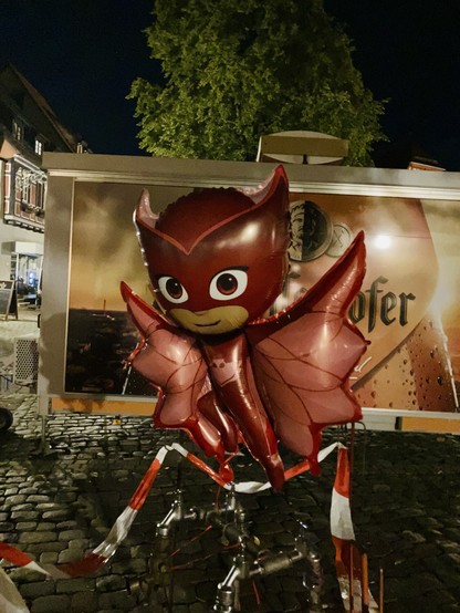 Ein roter Ballon in Form einer Zeichentrickfigur vor einem Bierverkäufer in der Nacht.

A red balloon shaped like a cartoon character in front of a beer vendor at night.