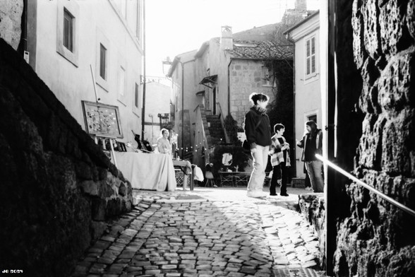 Schwarzweißfoto - Blick durch das Eingangstor in das Zentrum der kleinen historischen Stadt Calcata über Pflastersteine. Links und rechts Mauern und Häuser, in der Bildmitte ein kleiner Markt mit Kunstprodukten und verschiedene Leute - Aussteller und Besucher. Leica IIIg mit 50 mm Summitar auf Adox HR 50-Film.