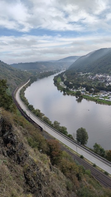 Blick von oben ins Tal. Die Mosel zieht sich vom rechten Rand in einer Kurve bis in die obere Bildmitte und verschwindet zwischen den Bergen. Bäume auf beiden Ufern, links Bundesstraße und Schienen, dann Felsen. Weiter hinten Hatzenport. Am rechten Ufer die Ortschaft Burgen mit Campingsplatz. Aufgelockerte Bewölkung mit blauen Flecken, in den dunkelgrünen Bergtälern hängt noch weißer Nebel.