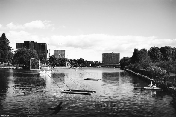 Schwarzweißfoto, aufgenommen von der Brücke am Ende des EUR-Sees im Süden von Rom. Im Vordergrund das Wasser des rechteckigen Sees mit Wasservögeln rechts auf einem Steg. In Bildmitte ein Tor für Kanupolo. Im Hintergrund hohe Geschäftsgebäude und Bäume. Leica R4 mit 35 mm Summicron auf Adox CMS II 20.