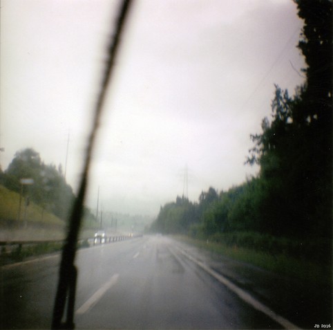 Farbfoto durch die Windschutzscheibe eines fahrenden Autos - unterwegs auf der Autobahn in der Schweiz im Regen. Der Scheibenwischer steht gerade fast senkrecht, auf der Fahrbahn sammelt sich Wasser. Links und rechts etwas verschwommene Landschaft, grauer Himmel mit Strommasten. Diana Mini auf Fuji C200-Film.