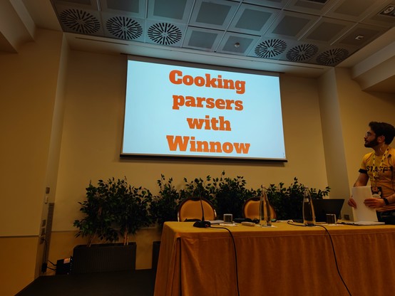 A screen showing the title slide "Cooking parsers with Winnie", in front of a yellow wall. Before the screen is a desk with two chairs and one of the organizers of RustLab in a yellow shirt.